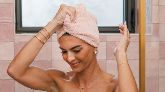 Woman wrapping wet hair in a pink towel turban in a tiled bathroom, smiling