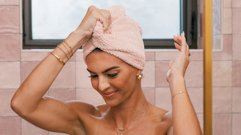 Woman wrapping wet hair in a pink towel turban in a tiled bathroom, smiling