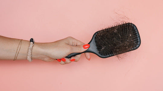 Hand holding a black paddle hairbrush clogged with shed hair against a pink background
