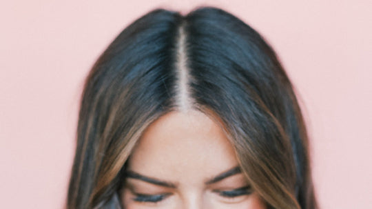 Woman with center-parted dark brown hair showing visible scalp at the crown, looking down against a pink background