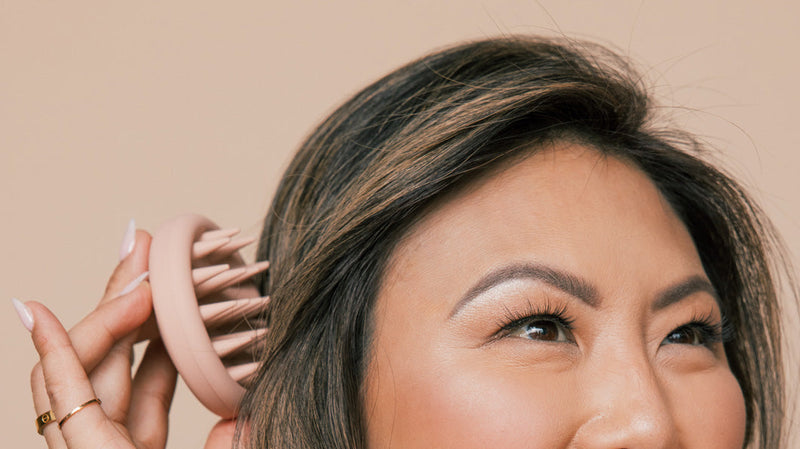 Woman using a pink silicone scalp brush at her hairline in a close-up against a beige background