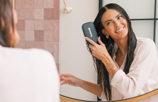 Smiling woman brushing wet hair while looking in a bathroom mirror with a black paddle brush.