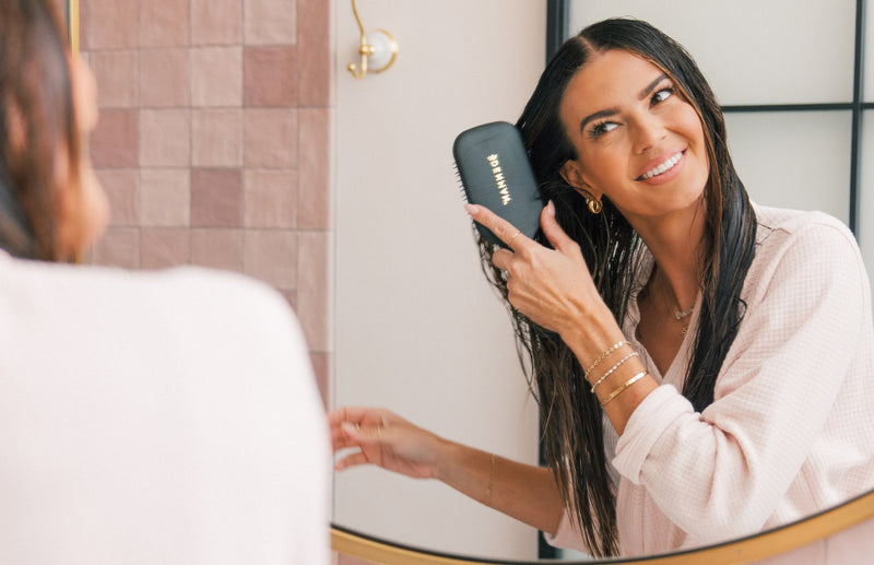 Smiling woman brushing wet hair with a black paddle brush in front of a bathroom mirror