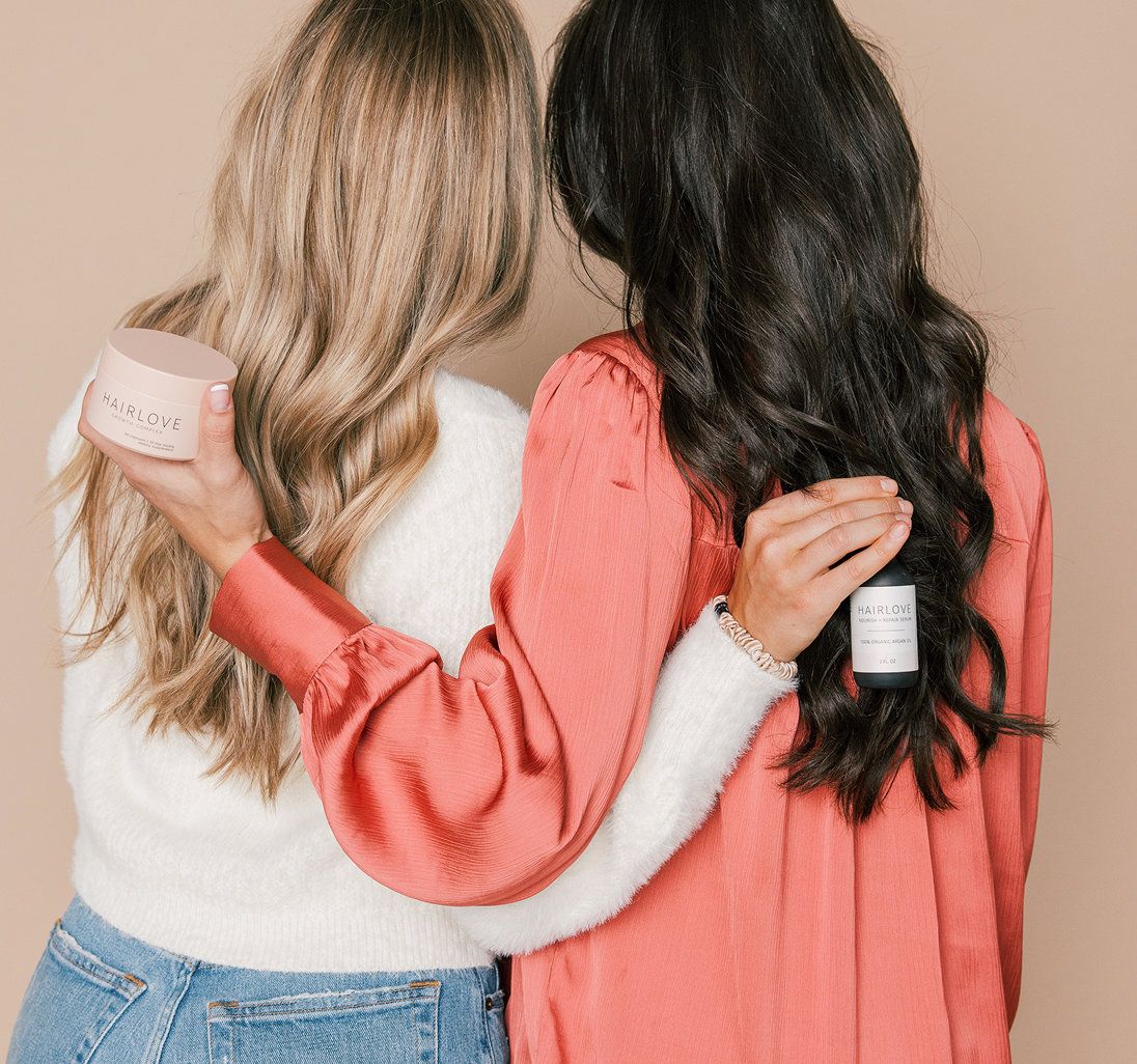Blonde and brunette women embrace, each holding a HAIRLOVE product jar behind their backs.