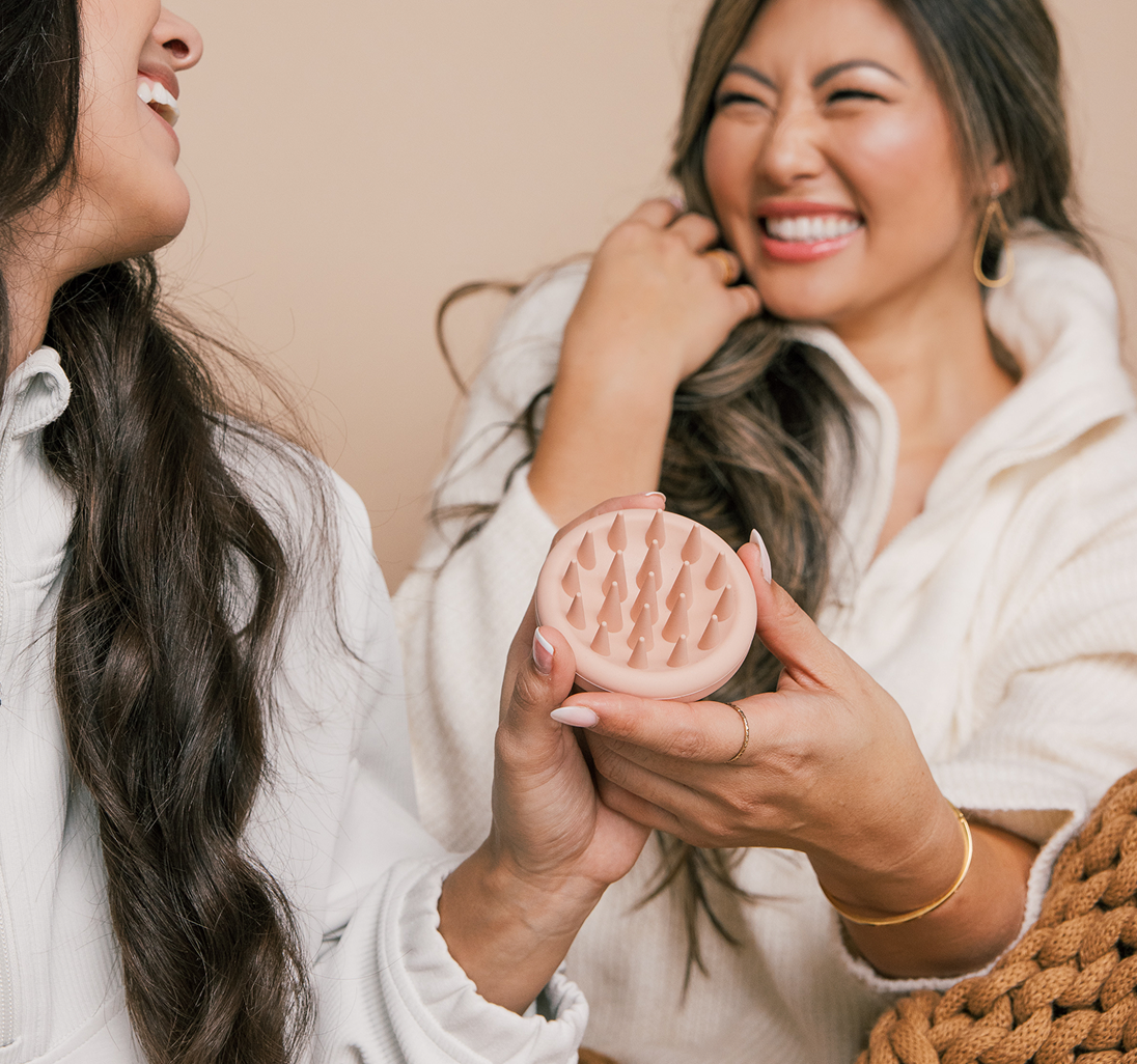 Two smiling women holding a pink silicone scalp massager with cone-shaped bristles