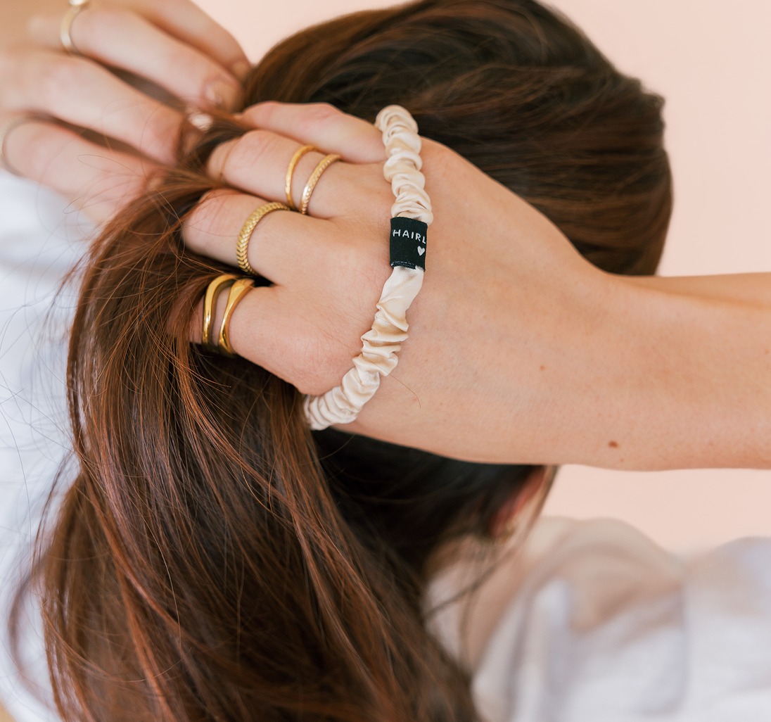 Hand gathering brown hair with a cream satin scrunchie labeled HAIRL and several thin gold rings