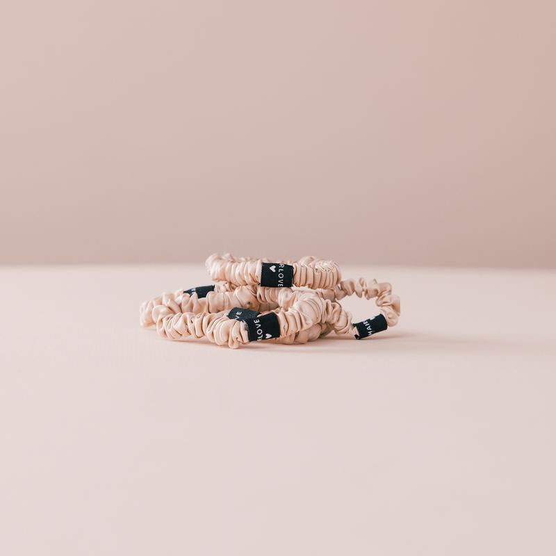 Beige scrunchie hair ties with black fabric tags that read LOVE, stacked on a pale pink surface