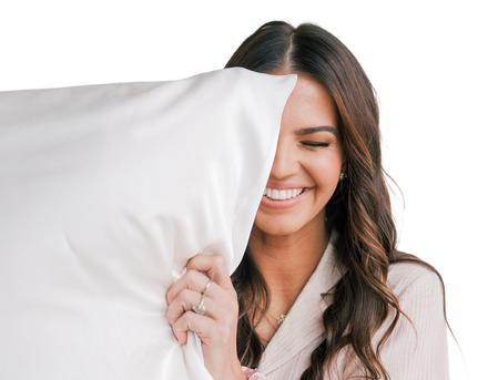 Woman smiling with eyes closed, holding a white pillow that covers one side of her face